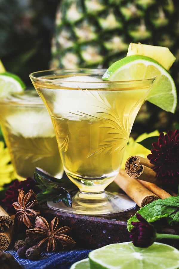 An iced glass of tepache sits surrounded with spices and adorned with lime and pineapple