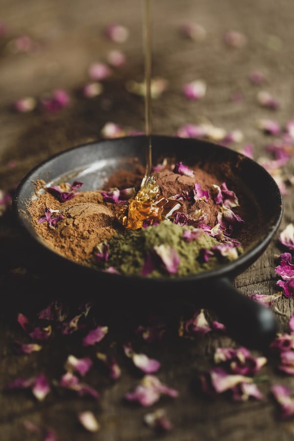 Bowl of herbal powders and rose petals with honey being poured into bowl. 