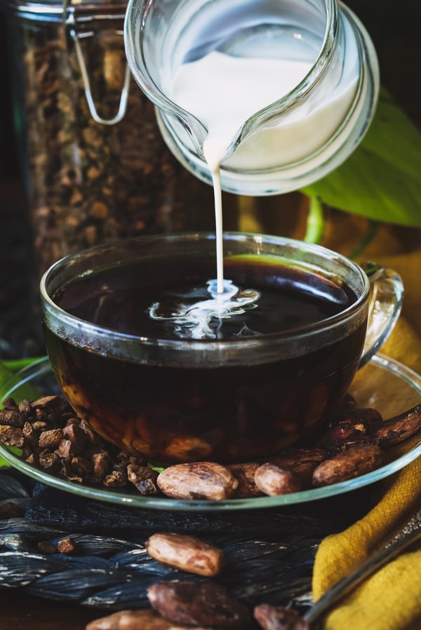 A glass mug of roasted cacao tea with cream being poured into it