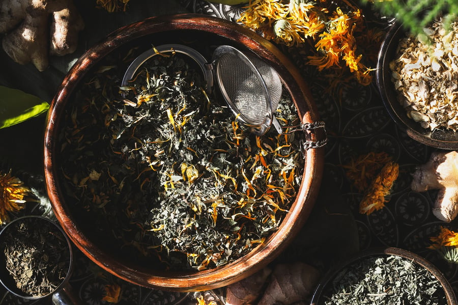 An overhead shot of herbal tea blend in wooden bowl with a tea ball sitting in it and calendula flowers nearby