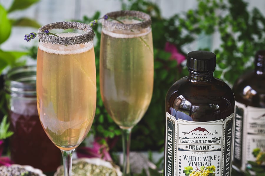 Two glasses of lavender yarrow oxymel mocktails near a bottle of white wine vinegar