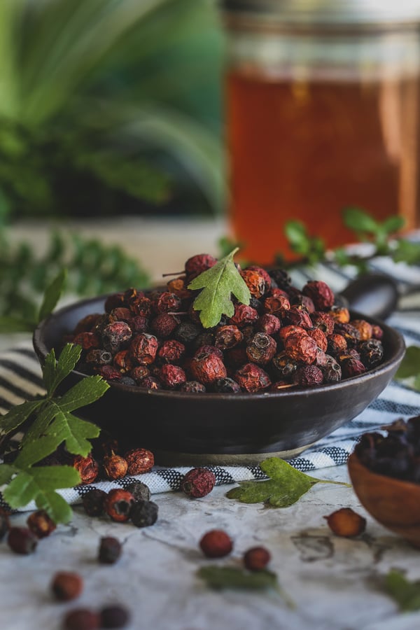 Bowl of dried hawthorn berries with fresh hawthorn leaves spread around. 