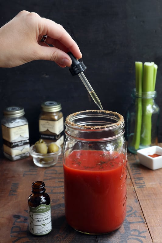Mason Jar of Bloody Mary with hand adding Horseradish Extract and garnishes in the background
