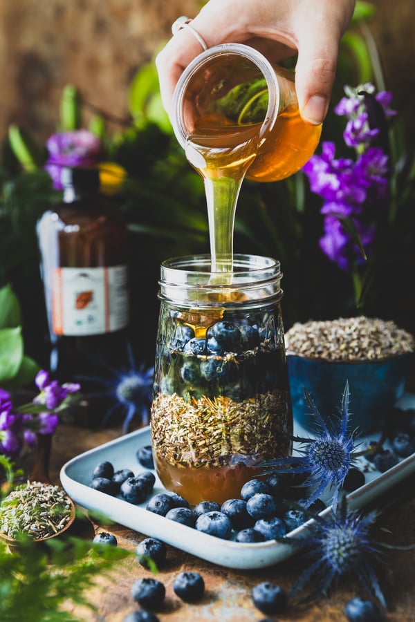 A honey is poured from the top right of the image into a glass jar filled with lavender flowers, blueberries, and apple cider vinegar. The jar is on a light blue tray surrounded by blueberries, a bowl of lavender, and fresh flowers.