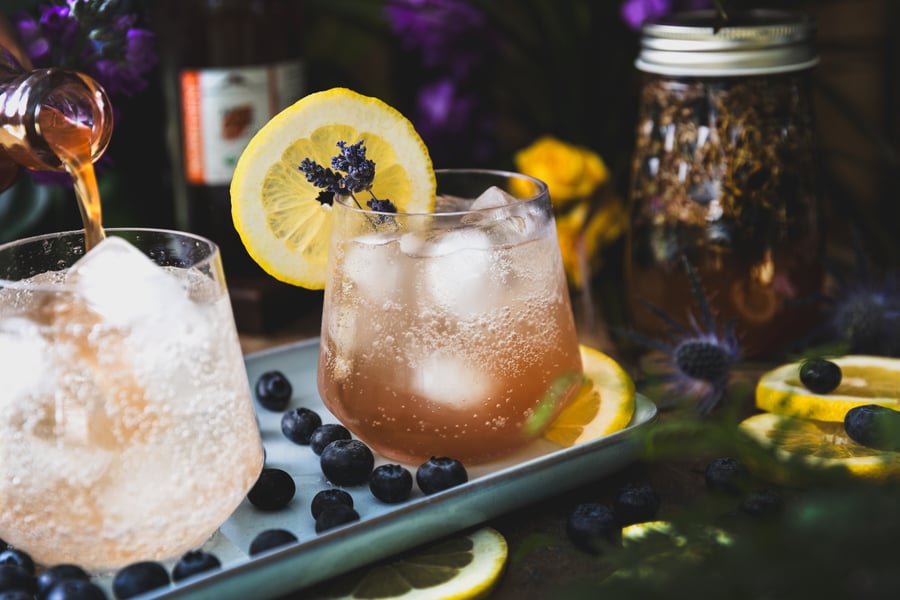 Two glasses of Blueberry Lavender Herbal Mocktails sit on a light blue tray surrounded by blueberries, lemons, a jar of blueberry lavender shrub, and cut flowers.