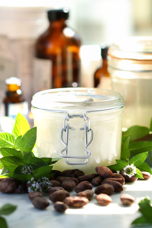 DIY Mint Chocolate Body Buttter in a jar surrounded by ingredients including mint leaves, cacao beans, organic carrier oils, etc.