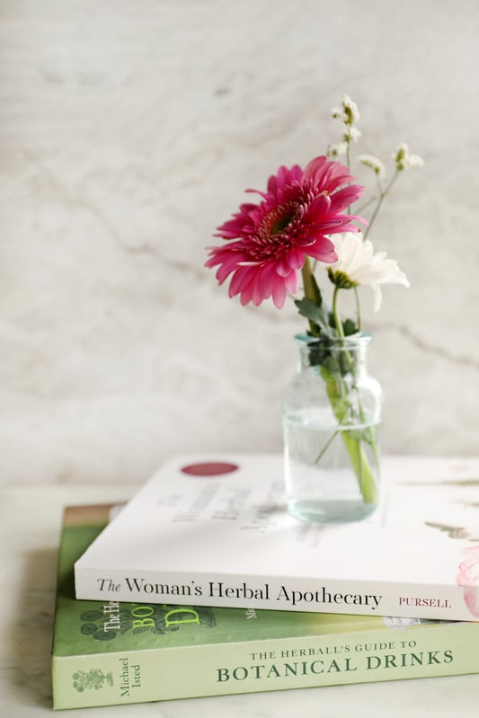 A short stack of books about herbalism with a glass jar full of bright, pink and white, spring flowers.