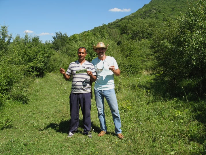 Mountain Rose Herbs procurement officer standing with farmer holding I Love Herbs bumper sticker in a field in Bulgaria