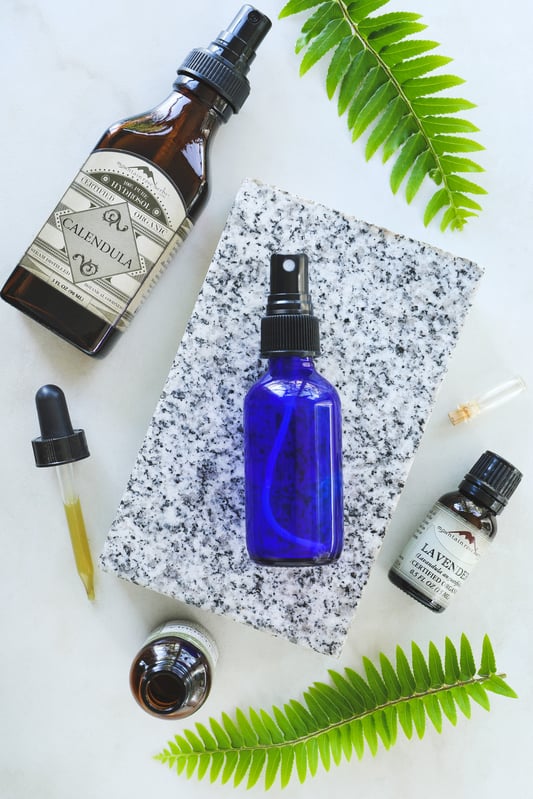 Materials and ingredients for calendula facial toner include a blue cobalt mister bottle, lavender essential oil, calendula hydrosol, and calendula extract. Arranged with fresh fern fronds and a slab of marble.