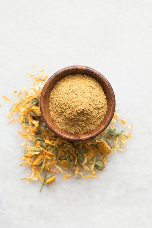 Wooden bowl full of organic calendula powder surrounded by calendula flowers on a white marble counter top