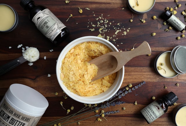 Wooden scoop in white bowl filled with carnauba wax with other ingredients for salve recipe making