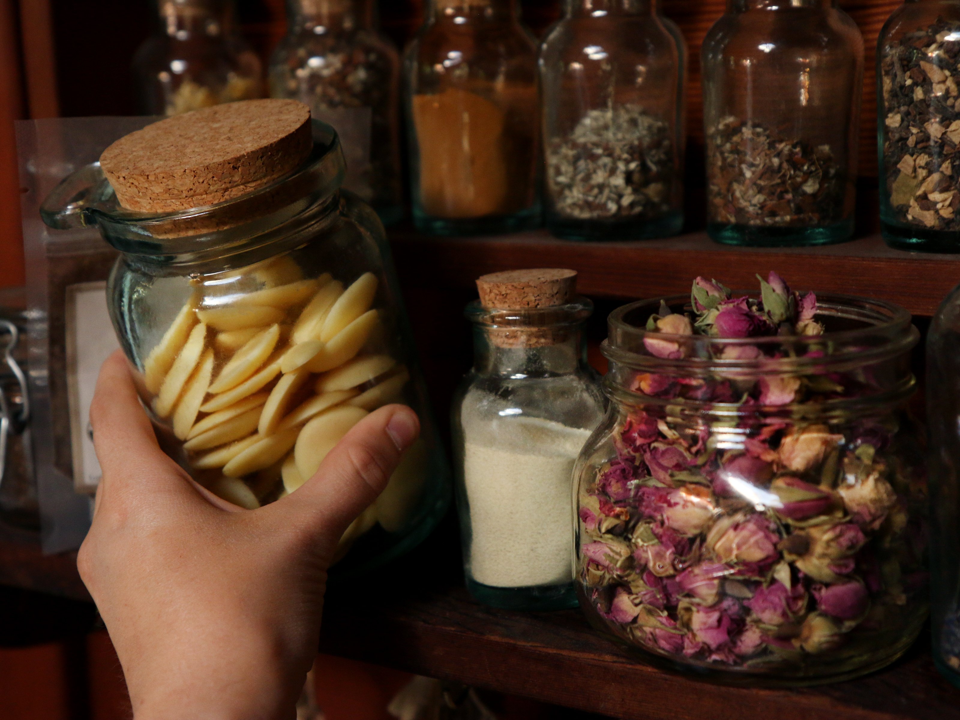 A shelf of herbs in glass jars with cork tops. Coconut butter wafers, rose buds, herbal powders, tea blends, and roots used to make herbal preparations. 