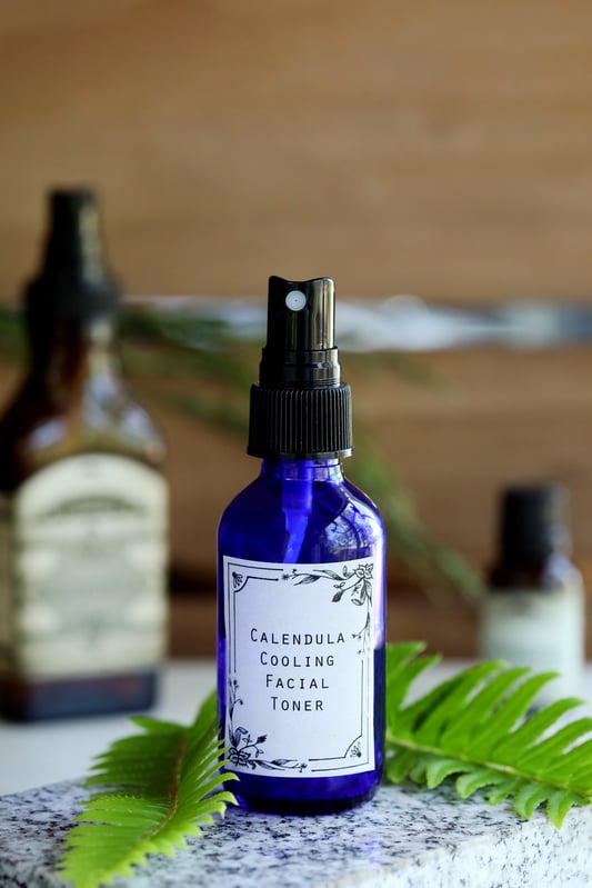 A blue cobalt spray bottle of calendula facial toner adorned with ferns atop a marble slab.