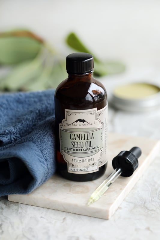 Bottle of Camellia Seed Oil displayed with a dropper, bright green plant and a crisp blue towel. 