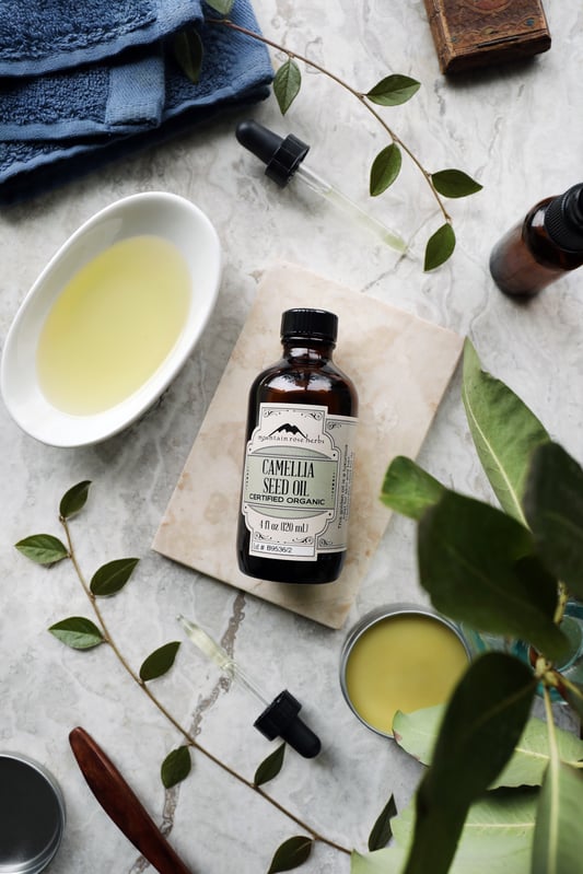 Bottle of Camellia Seed Oil displayed with a bowl of the oil, fresh plants and a towel. 