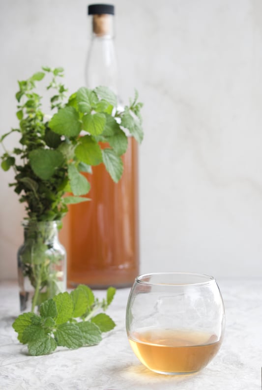 Clear globe glass holding Carmelite water near fresh lemon balm with bottle of amber liquid behind