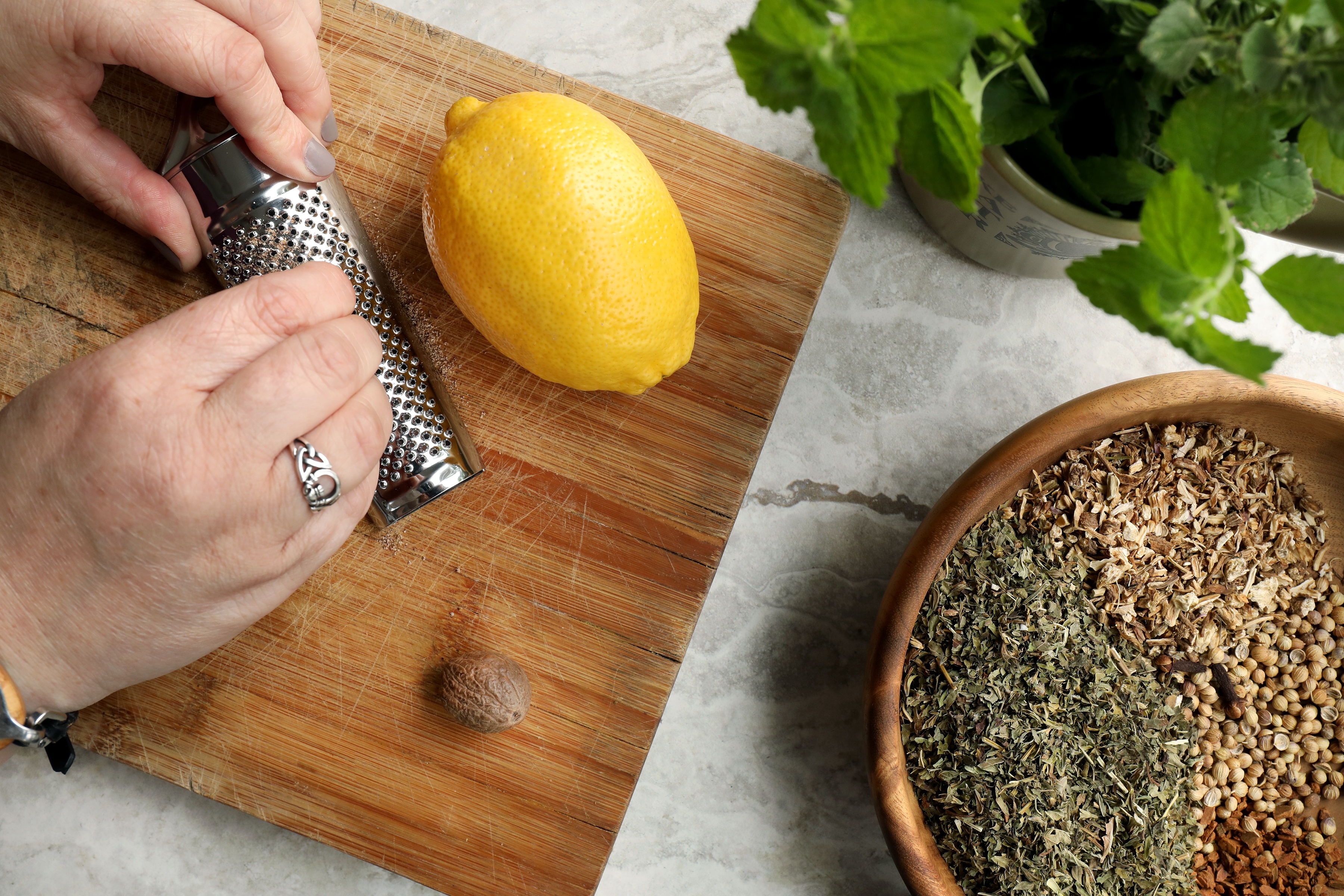 Hands grating nutmeg on wooden cutting board near lemon balm and lemon and dried herbs and spices.