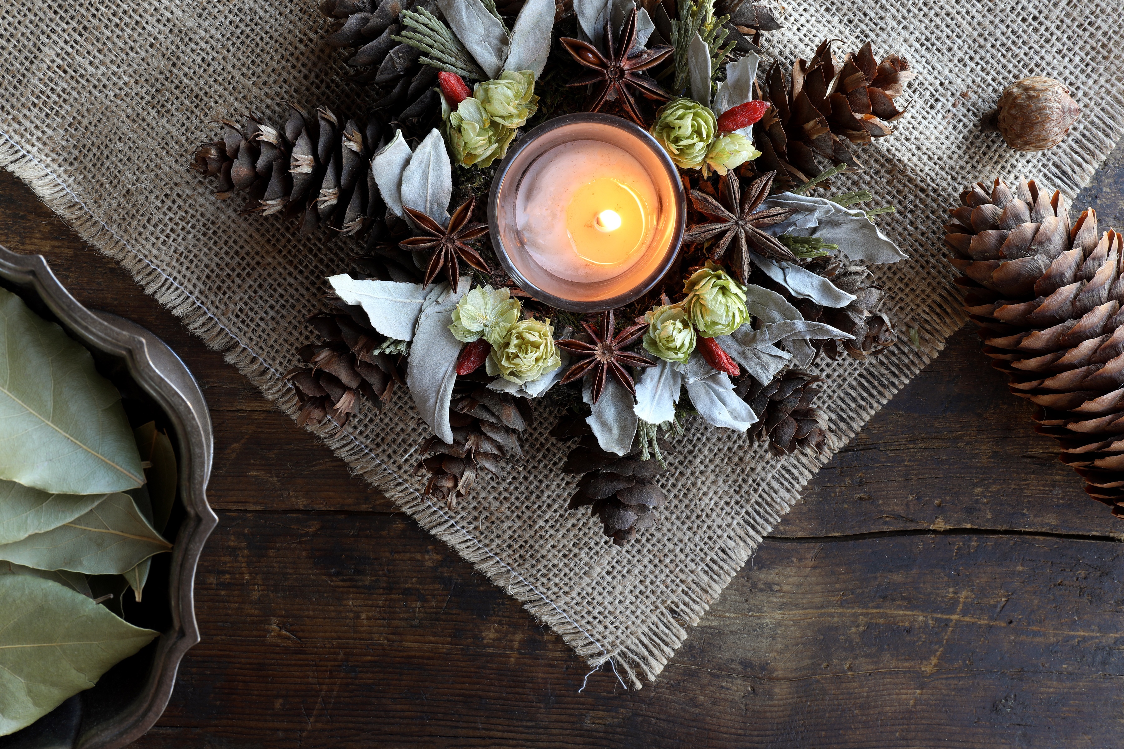 Center piece with holiday foliage decor and candle burning in middle , displayed on rustic wooden table and burlap