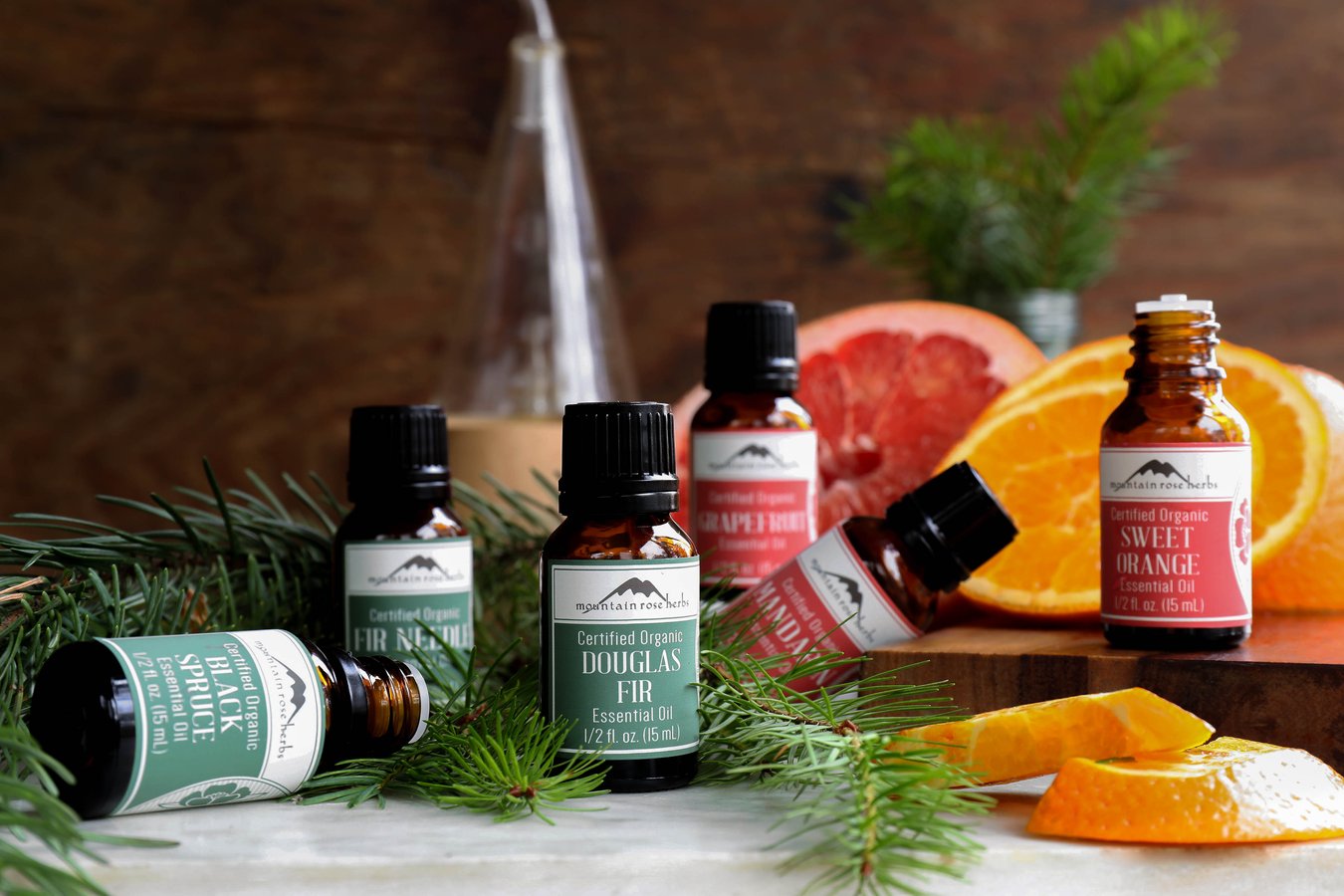 Black Spruce, Fir Needle, Douglas Fir, Grapefruit, Mandarin, and Sweet Orange essential oils surrounded by fir boughs and citrus.