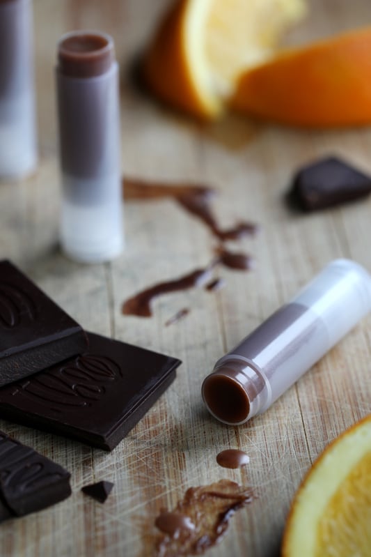 Dark colored lib balm in clear tubes with pieces of chocolate, fresh orange and drippings of chocolate. 
