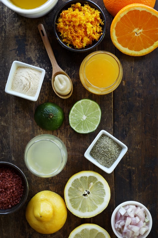 Ingredients for a citrus dressing are arranged on a rustic wooden tabletop. Ingredients include fresh lemon and orange, minced shallot, lime, vinegar, salt and orange rind.