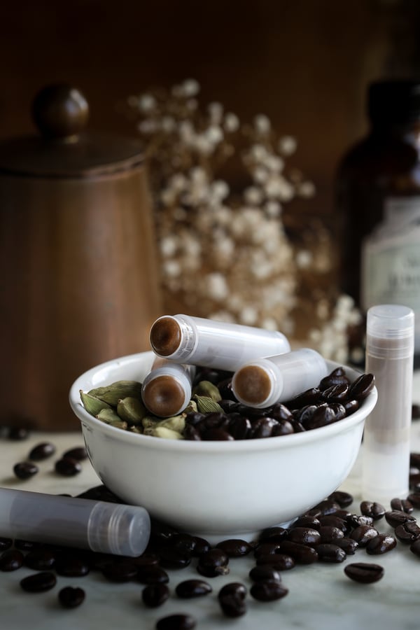 A bowl full of coffee beans and cardamom pods with tubes of coffee cardamom lip balm on top.