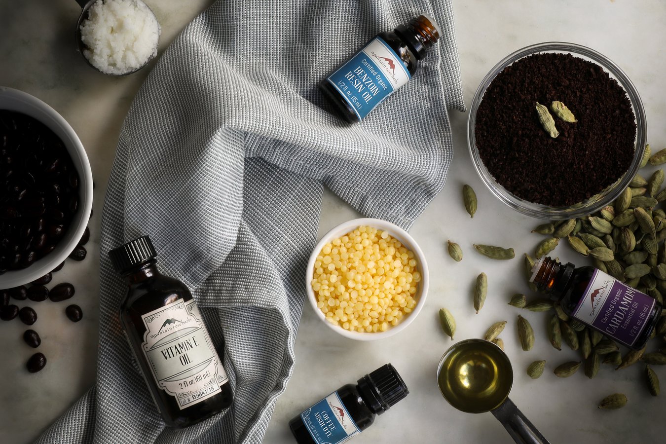 Ingredients to make uplifting coffee cardamom lip balm laid out on a table, including coffee beans, cardamom pods, vitamin e oil, essential oils and beeswax pastilles.