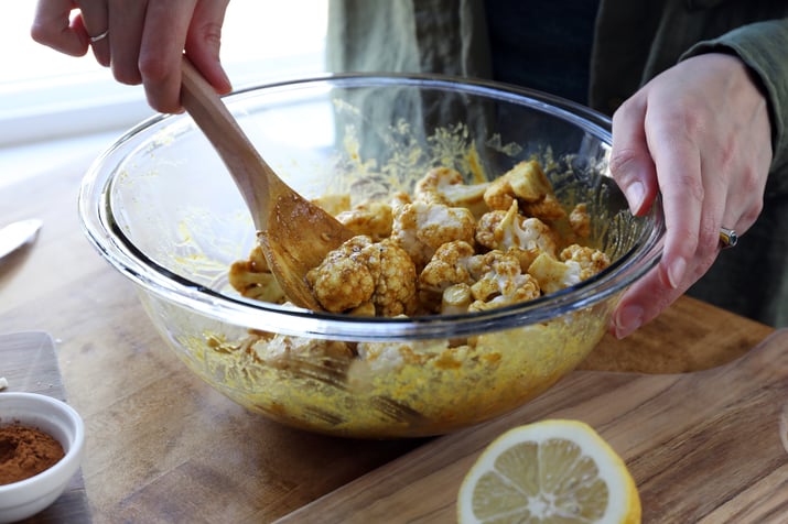Hands holding wooden spoon and glass mixing bowl stirring cauliflower into curry blend