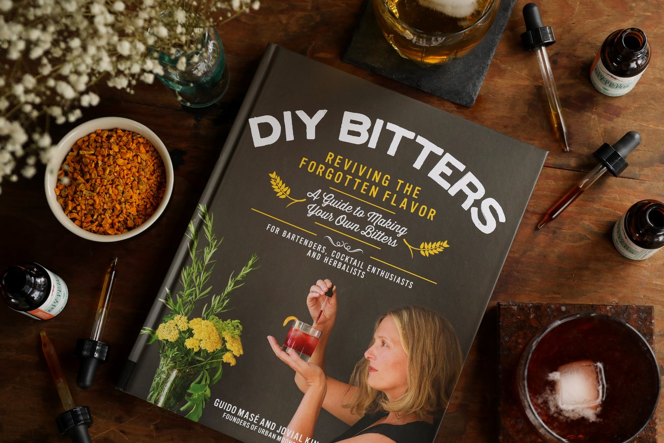 Hardback copy of book DIY Bitters: Reviving the Forgotten Flavor, by Guido Masé and Jovial King on a table surrounded by bottles of bitters, a cocktail made with bitters, and pipettes for measuring digestive bitters.