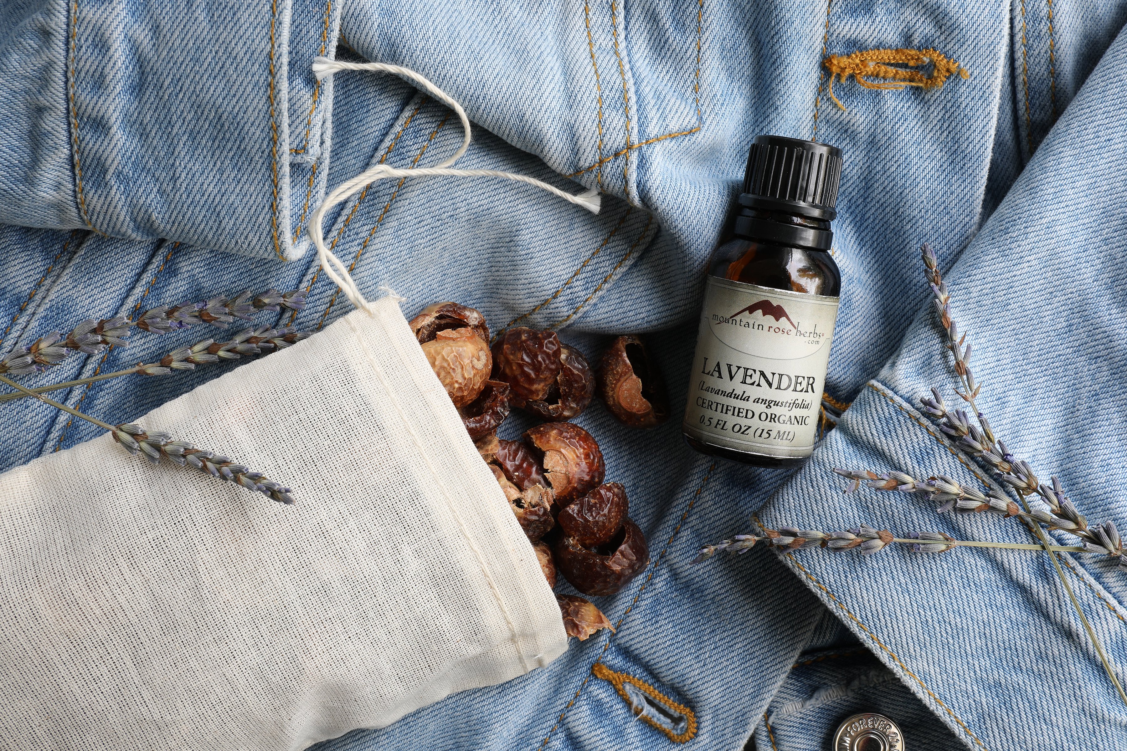 Bag of soap nuts spilling out of cotton bag with bottle of essential oil sitting on denim fabric. 