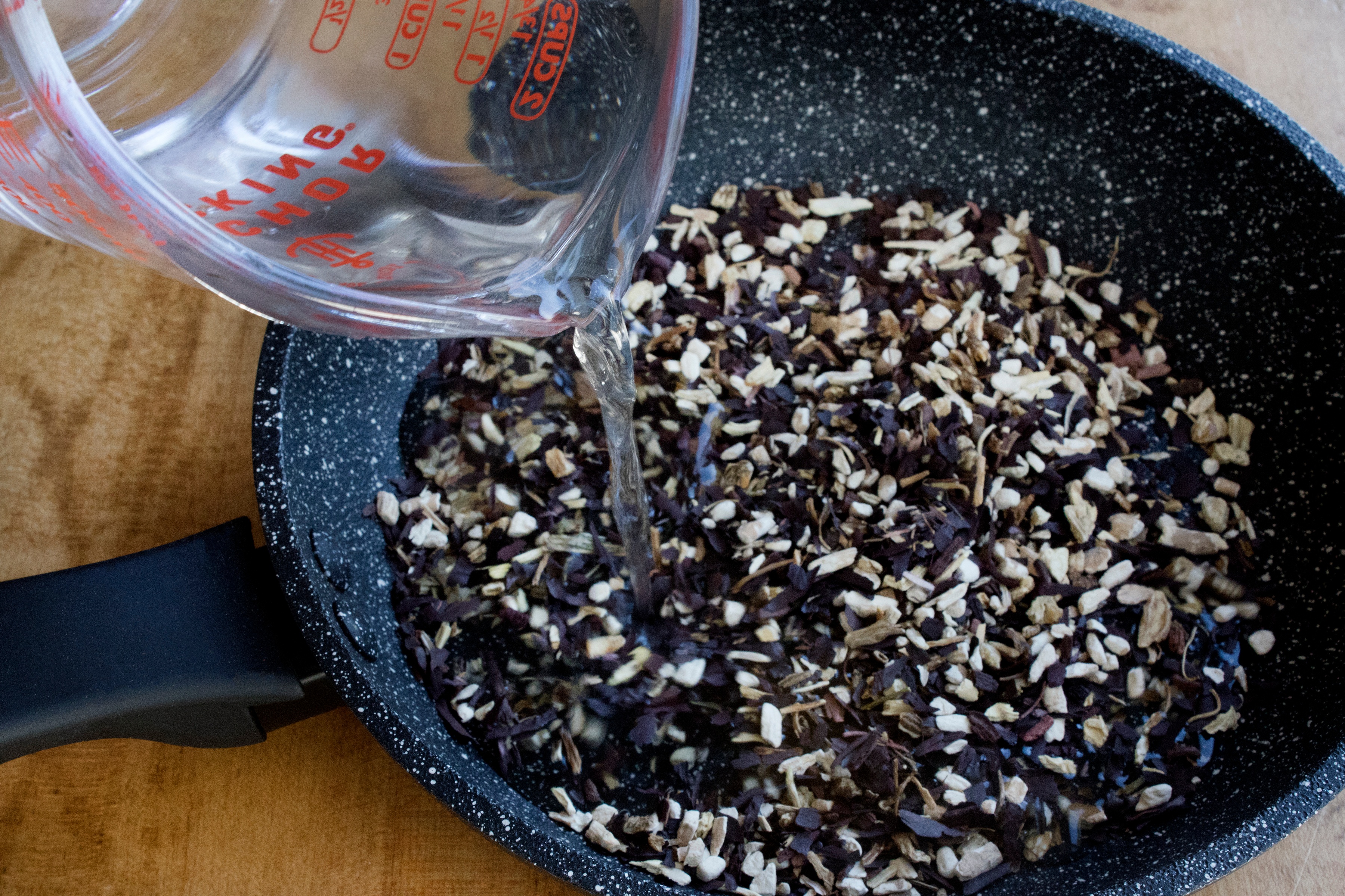 Dried cut roots in for sauté pan with measuring cup pouring water. 