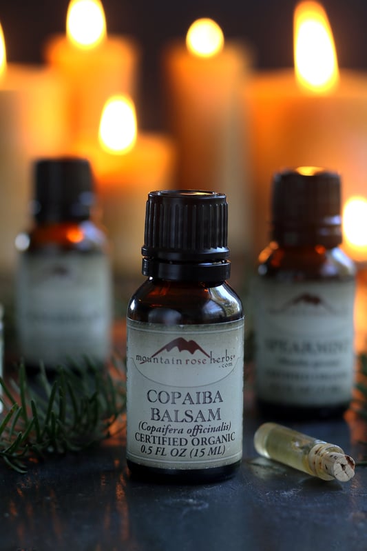 Several essential oils to make holiday blends for your home aromatherapy diffuser. Beeswax candles are lit in the background and copaiba balsam essential oil is featured. 