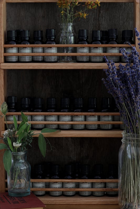 Essential oils stored on a wooden shelf in bottles indoors surrounded by dried herbs in mason jars