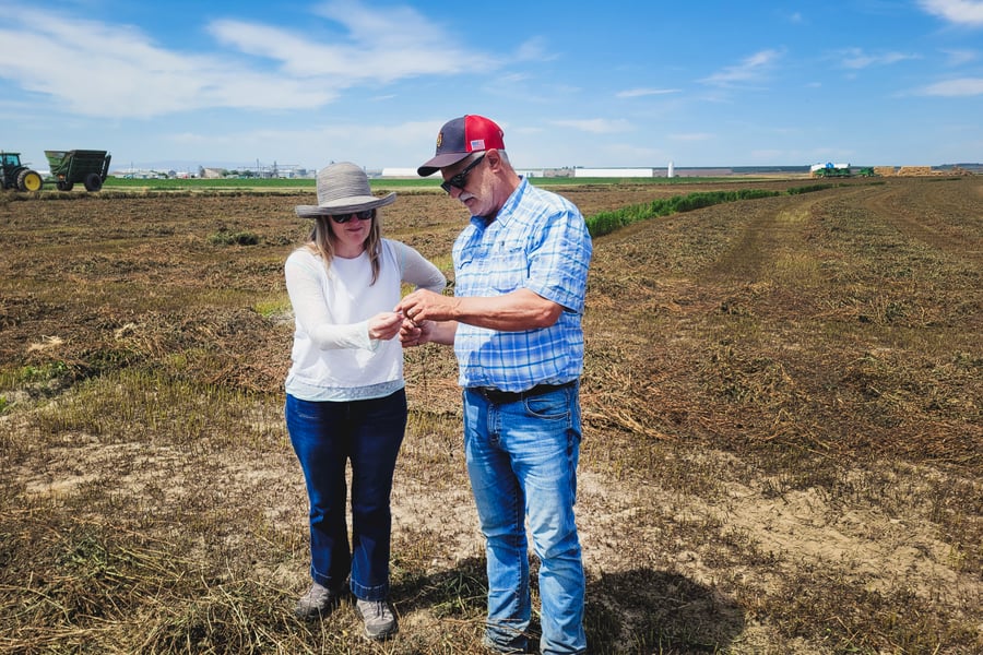 Cameron Stearns, Mountain Rose Herbs CEO, talks with a farmer about his crops. They are standing in farm fields with some buildings and a tractor behind them.