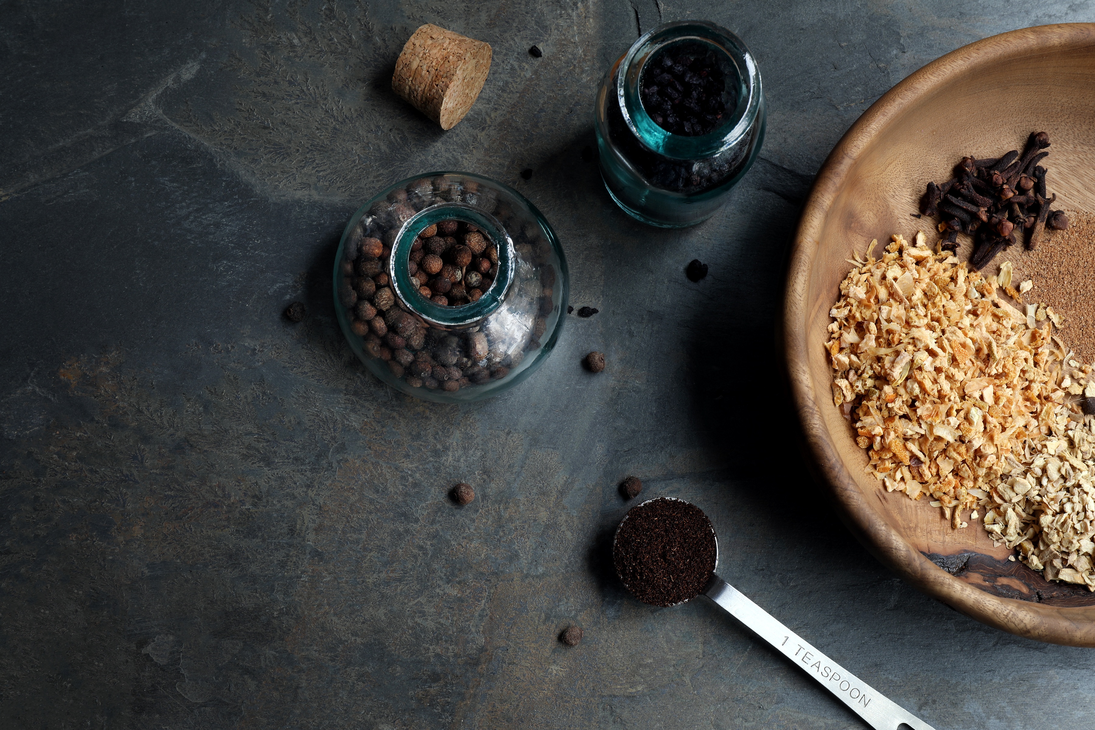 Dried herbs and spices spread out on plate, measuring spoons and glass jars of peppercorns. 