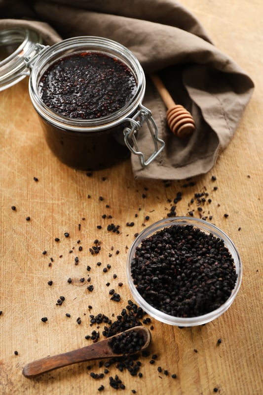 Ingredients for making elderberry vinegar are scattered on a table top. Finished vinegar rests in a large storage jar next to a wooden honey dipper and brown cloth towel, while freshly dried elderberries rest in a glass bowl next to a wooden spoon.
