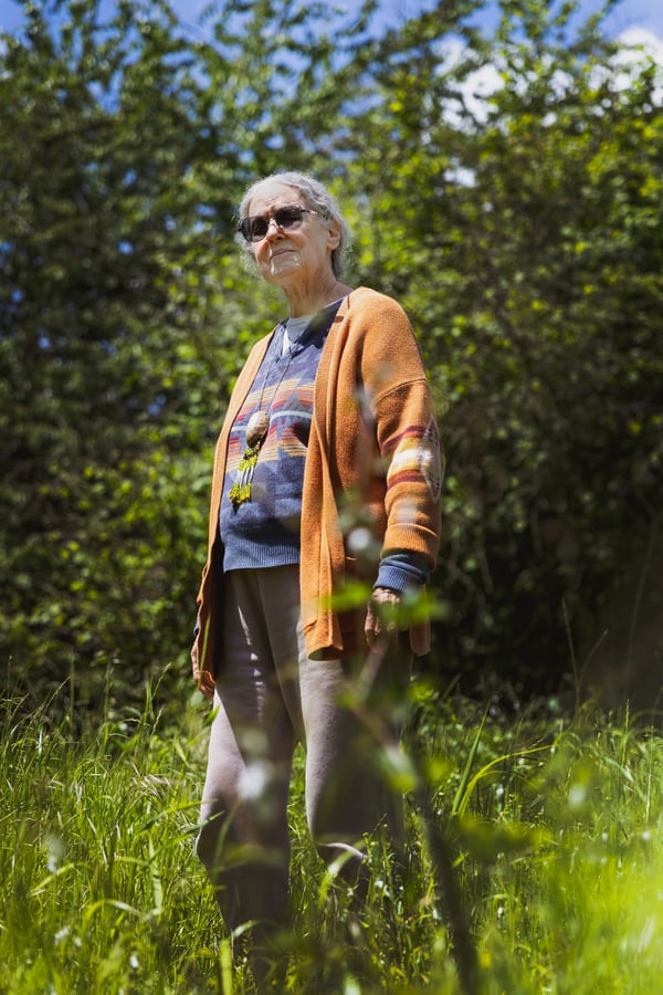 Rose High Bear, founder of Elderberry Wisdom Farm, stands on her property surrounded by the native plants that they steward in Salem, OR. Rose High Bear is wearing a patterned blue shirt with a bright orange sweater. She has gray hear, dark sunglasses on, and stands tall in a field of high grass.
