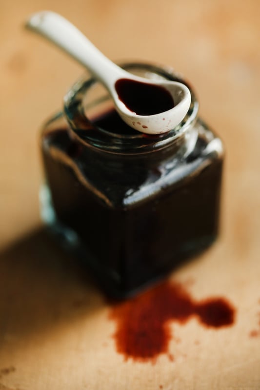 A white spoon full of dark, elderberry vinegar rests on the mouth of a clear jar full of dark, syrupy liquid. Several drops of dark red vinegar drips onto a wood surface.