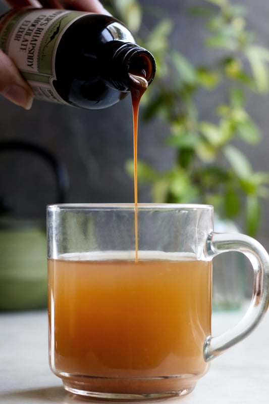 An herbal chocolate syrup is added to a warm beverage in a clear glass mug. Herbal syrups are used for natural wellness.