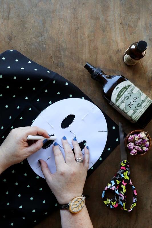 A DIY enthusiast is tracing a face mask template onto patterned fabric to make a homemade facial mask for skin care. Using Rose hydrosol and organic rose buds and organic rosehip seed oil.