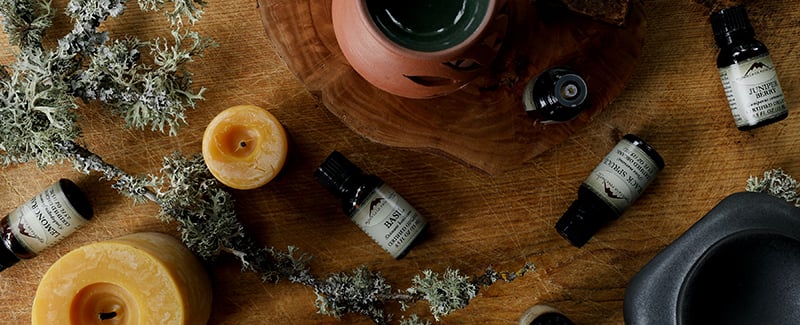 A collection of essential oils arranged on a wooden table with beeswax candles and terra cotta diffusers. A branch from an oak tree is covered in lichen. 