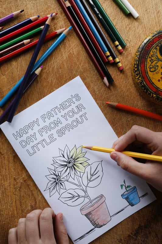Coloring in a Father's Day card featuring a grown flower and a sprout in pots. Colored pencils are scattered around the card on a wooden table. 