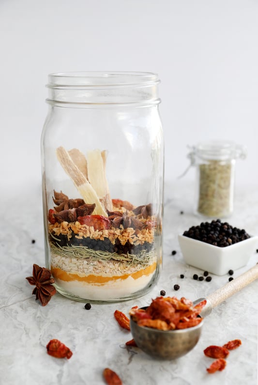 Clear quart canning jar with colorful layered spices and other botanicals surrounding it. 