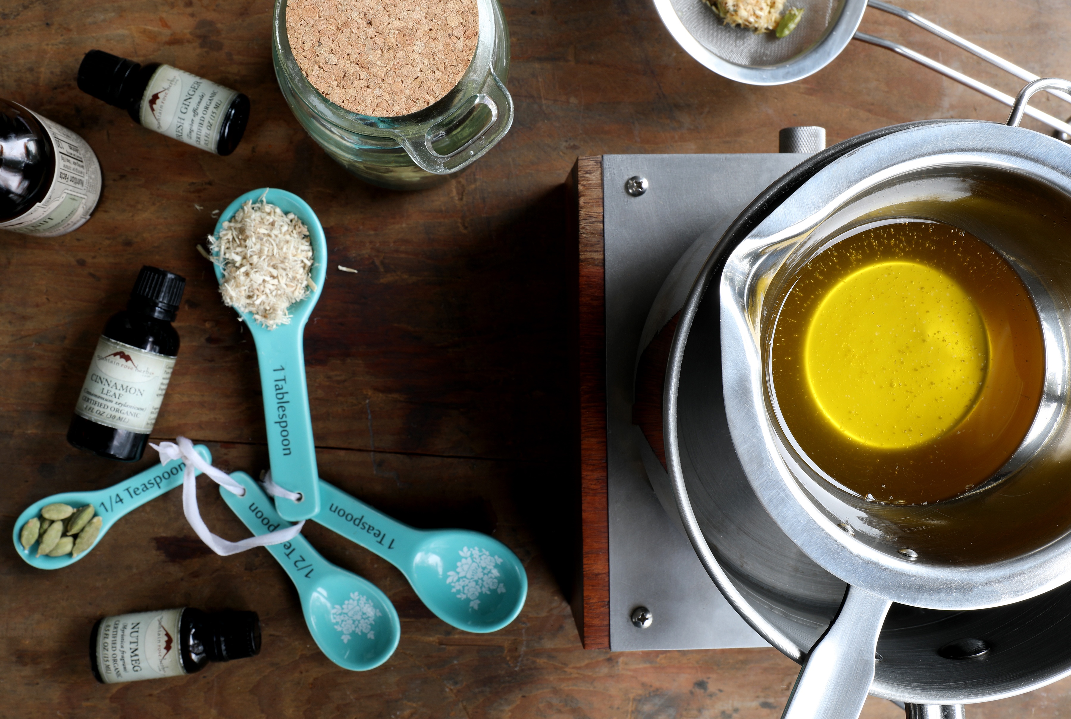 Double boiler inside of pot containing golden colored oil.  Displayed measuring spoons with ingredients in them.  Setup for making lip balm at home.