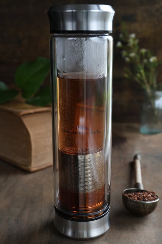 Glass walled tea-to-go travel cup filled with amber colored tea with an antique book and plant in background. 