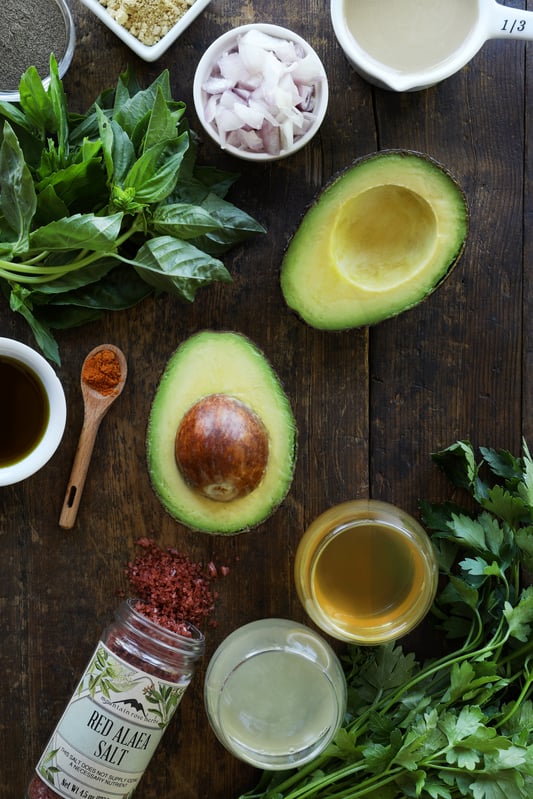 Fresh, colorful ingredients are arranged together including sliced avocado, minced shallots, basil, parsley, apple cider vinegar, red Alaea salt, and lemon juice, on a rustic wood table top.