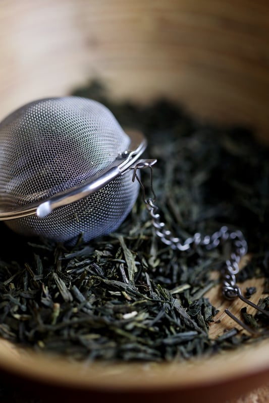 Dried green tea leaves with a mesh steel tea ball in a wooden bowl