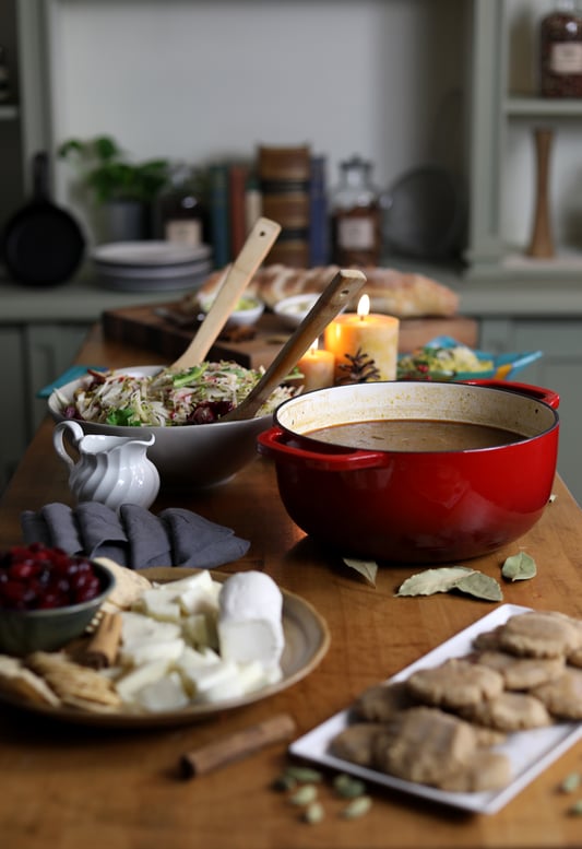 Table with food for group potluck including pot of soup, plates with chees and cookies, and greens and bread and candles