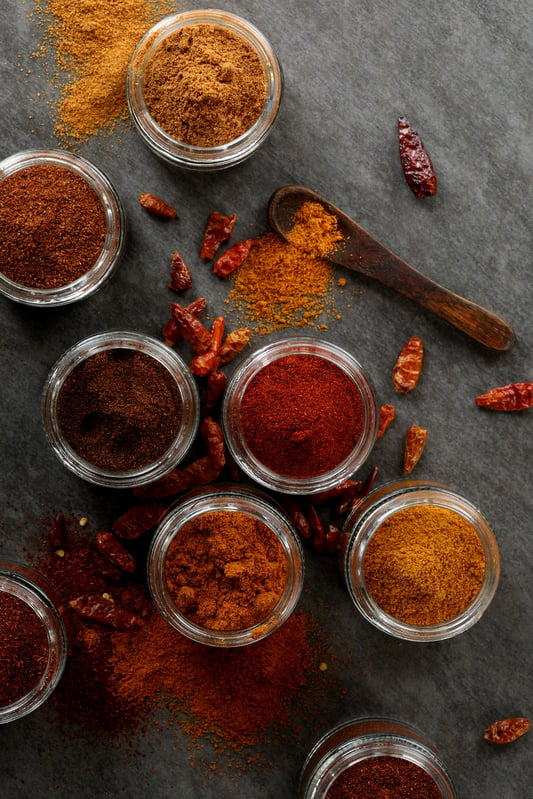 A variety of chili powders arranged in clear glass jars. Paprika, cayenne, habanero, bird's eye chili, and chipotle powders in various shades of red and brown. 
