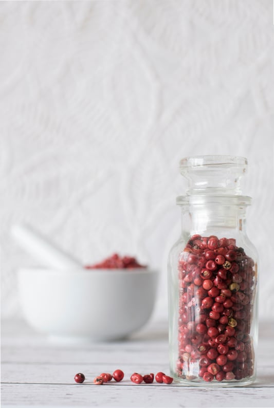 Different types of pink peppercorns in glass jar on counter with a mortar and pestle in the background